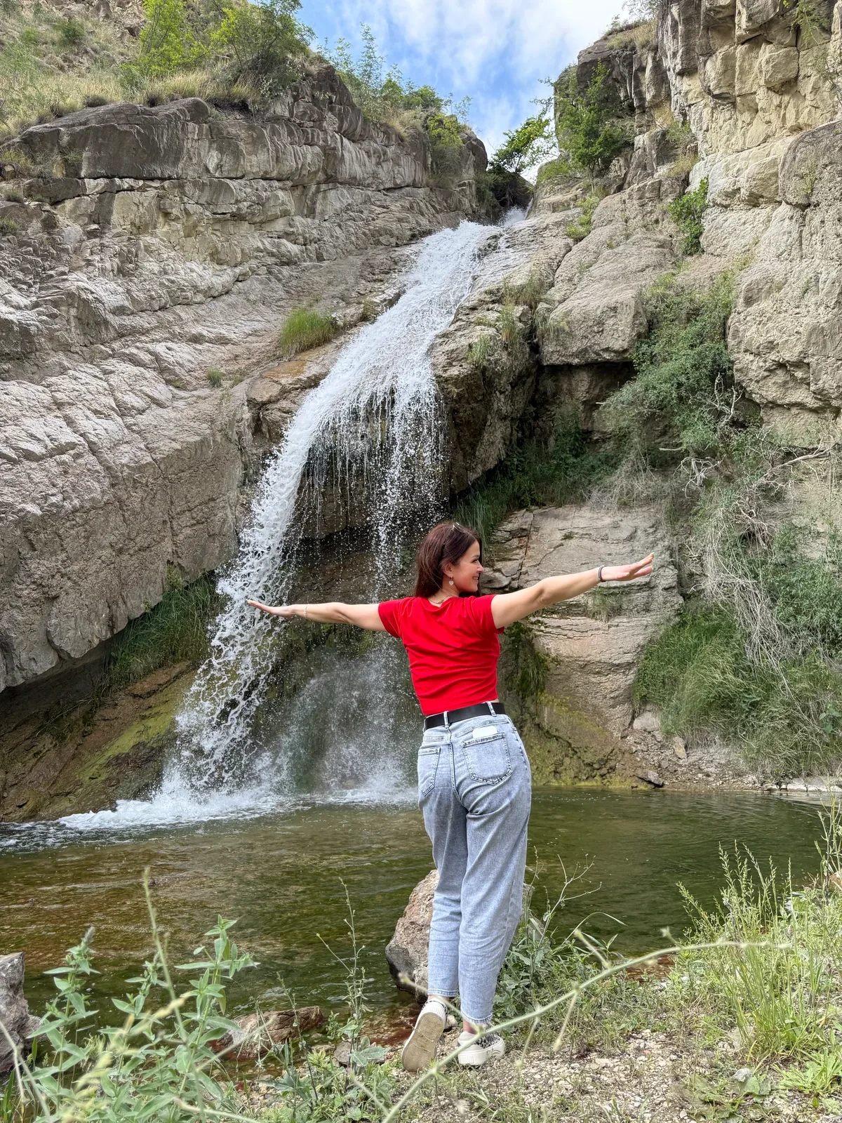 Человек в красной футболке перед небольшим каскадным водопадом и скалами